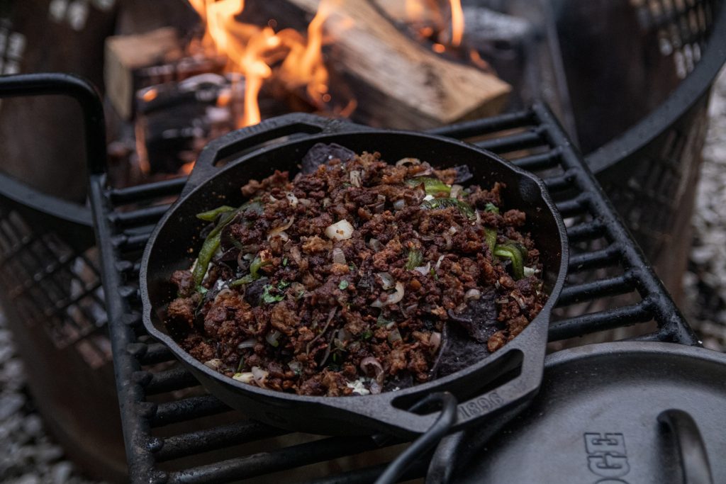 A Lodge cast iron pan full with Beyond Meat beef crumbles and nachos sits atop a fire pit grate with a campfire in the background.