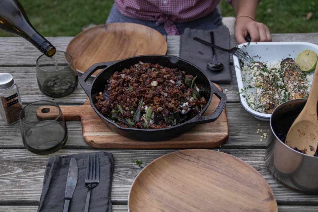 A woman pours herself a glass of white wine, ready to dig into the food on the table: black beans, Mexican street corn, and nachos.