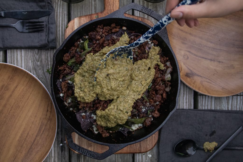 Someone spoons salsa verde onto a Lodge cast iron pan full of nachos. 