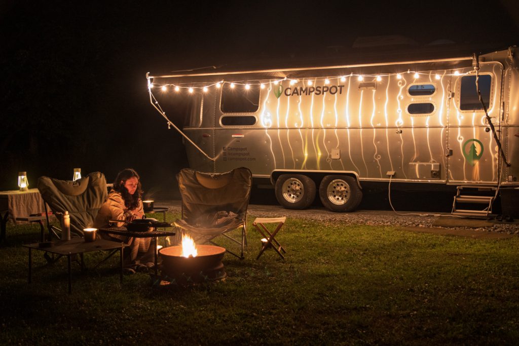 A woman keeps warm around the campfire with an Airstream trailer in the background lit with string lights.