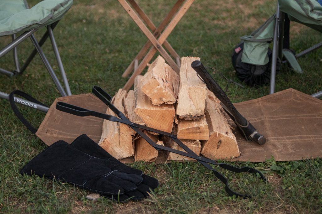 A pile of campfire wood on the ground with an axe, log grabber, and heat resistant gloves laying on it.