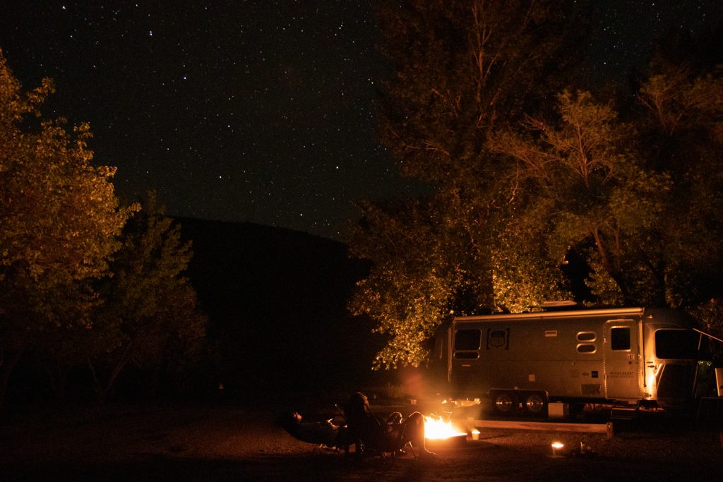 A couple sit around their campfire and Airstream trailer looking up at the starts at Capitol Reef National Park in Utah.