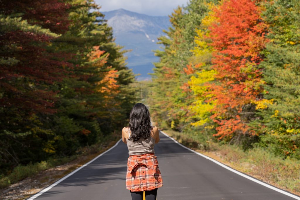 A woman stands in the middle of the road at Baxter State Park in Maine, taking a picture of Mount Katahdin and surrounded by fall foliage.