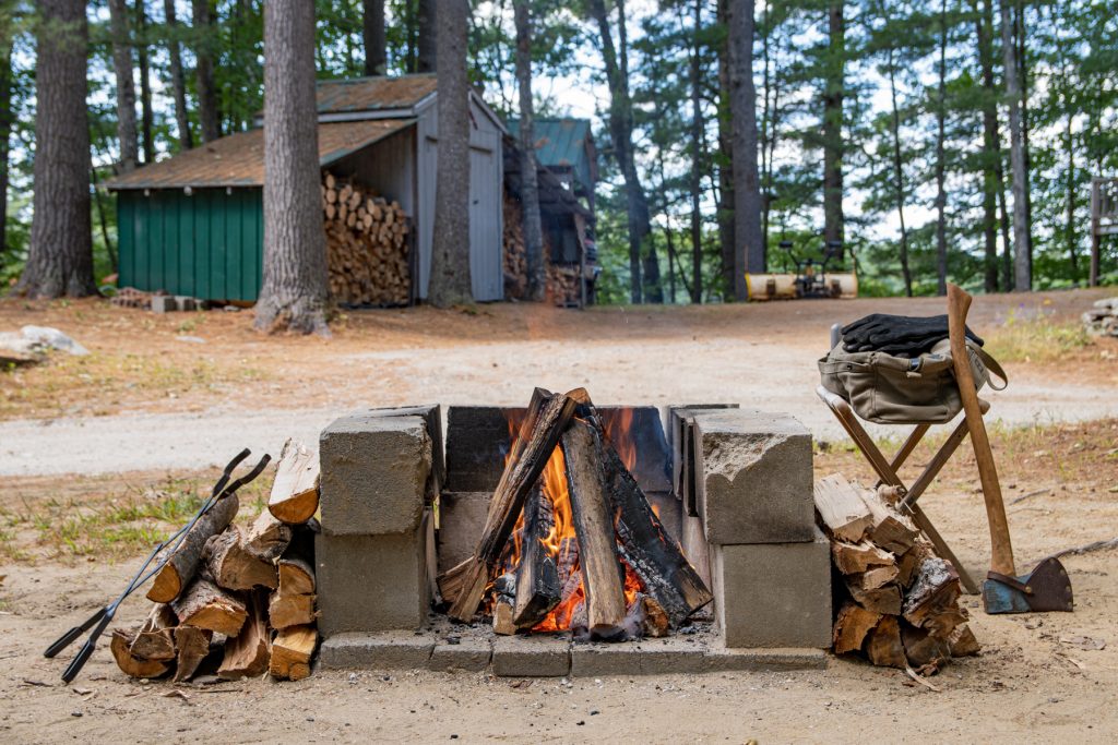 A roaring campfire surrounded by campfire wood, an axe, and a cabin full of campfire wood in the background.