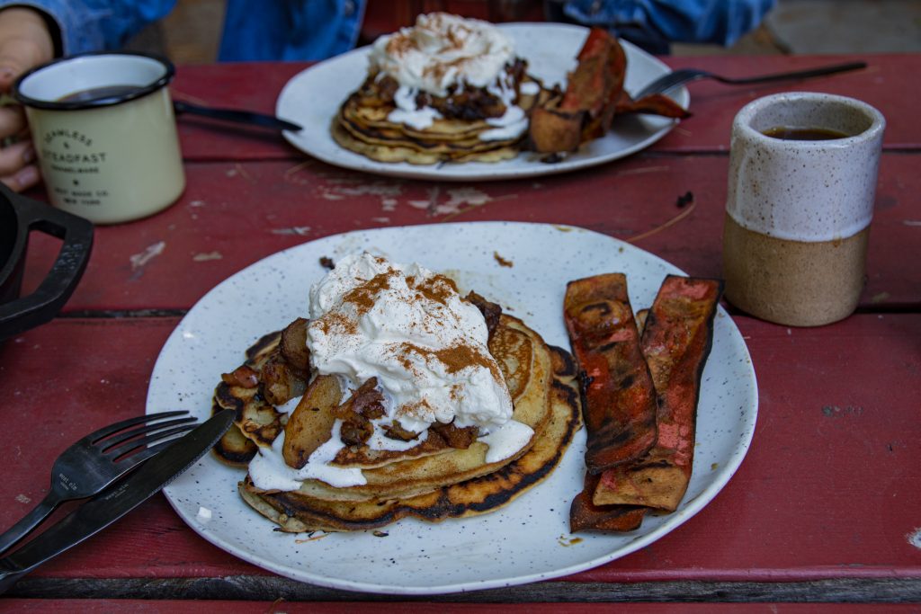 Two plates full of apple bourbon pancakes and bacon with mugs of coffee.