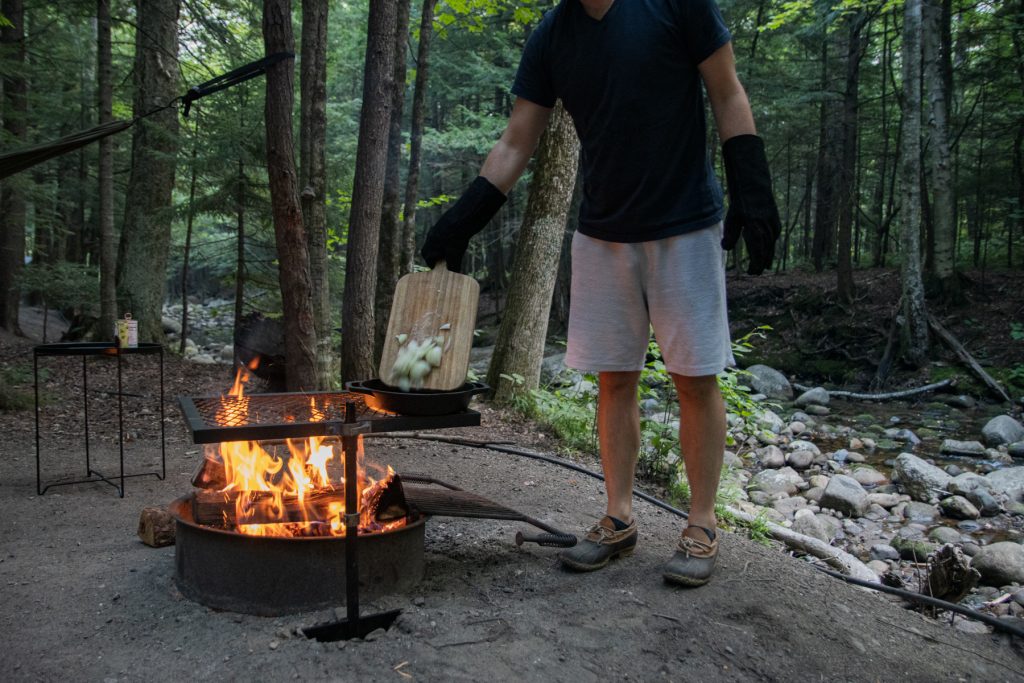 A man pours sliced onions into a Lodge cast iron pan sitting on top of a campfire grill with a roaring fire underneath.