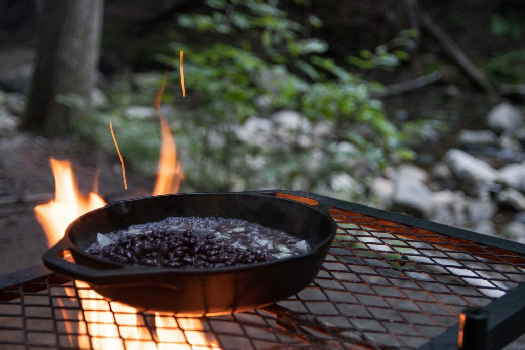 A Lodge cast iron pan full with black beans and onions sitting on top of a campfire grill with flames coming up from beneath.