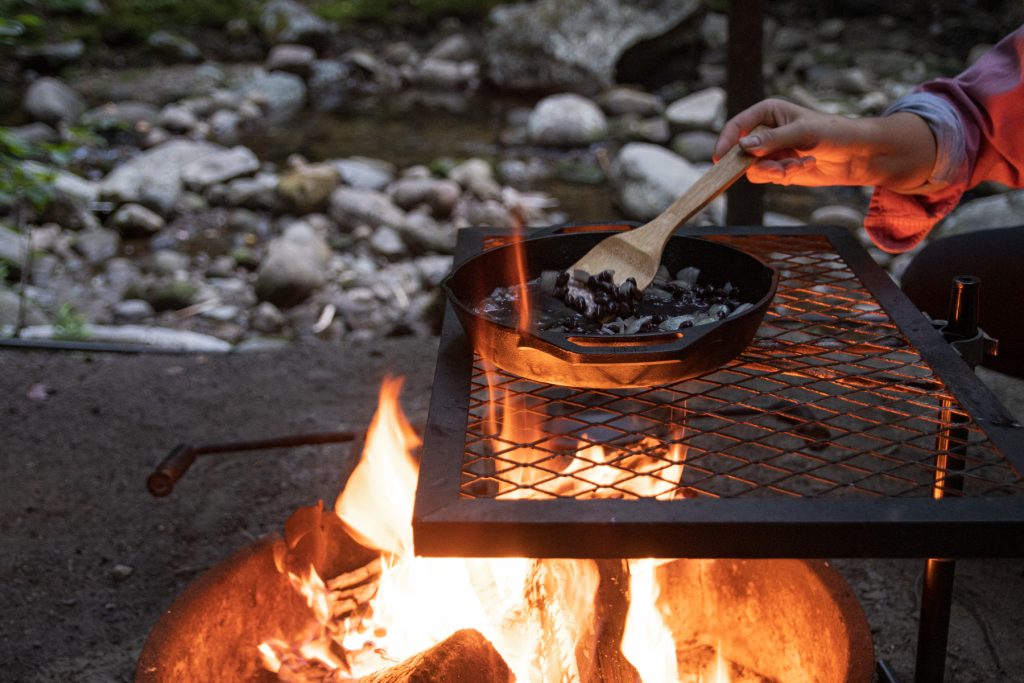 A person carefully stirs the Lodge cast iron pan of black beans with campfire flames coming up through the campfire grill.