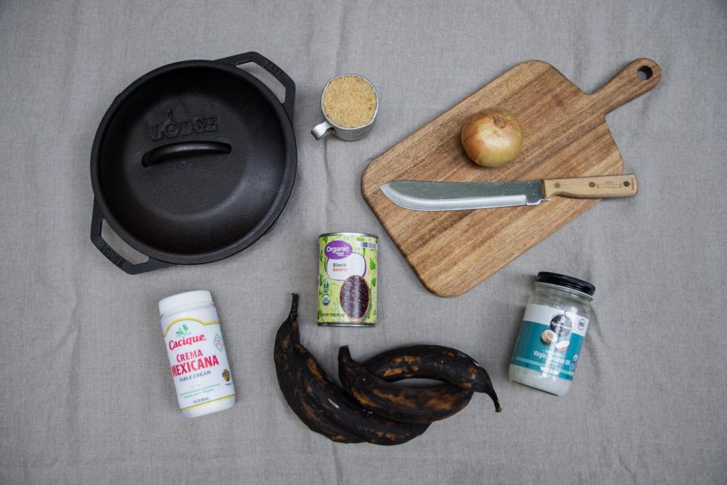 A top-down image of a Lodge cast iron pan, wooden cutting board, crema Mexicana, coconut oil, plantains, a can of black beans, onion, and cup of rice.