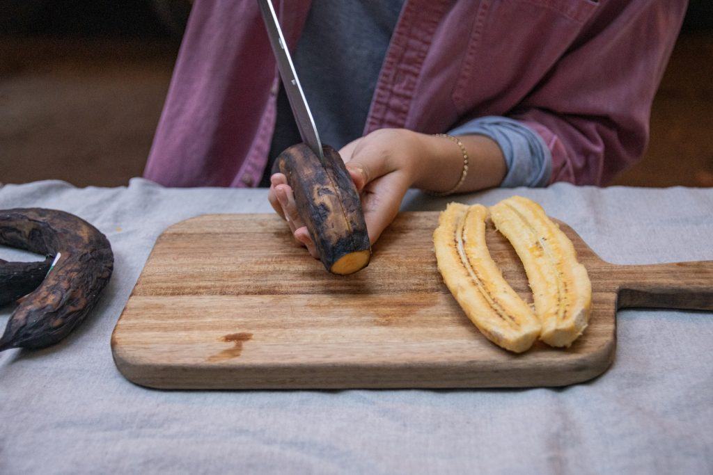 A person slices the peel of a ripe plantain length-wise on top of a wooden cutting board.