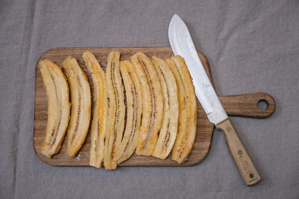 A top-down image of plantain halves sitting on top of a wooden cutting board with a knife next to them.