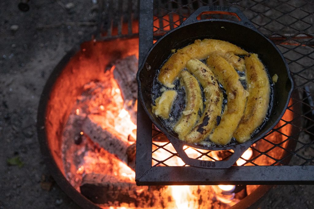 A top-down image of the plantain halves frying in coconut oil within the Lodge cast iron pan, sitting on top of the campfire grill grate and with glowing embers below.