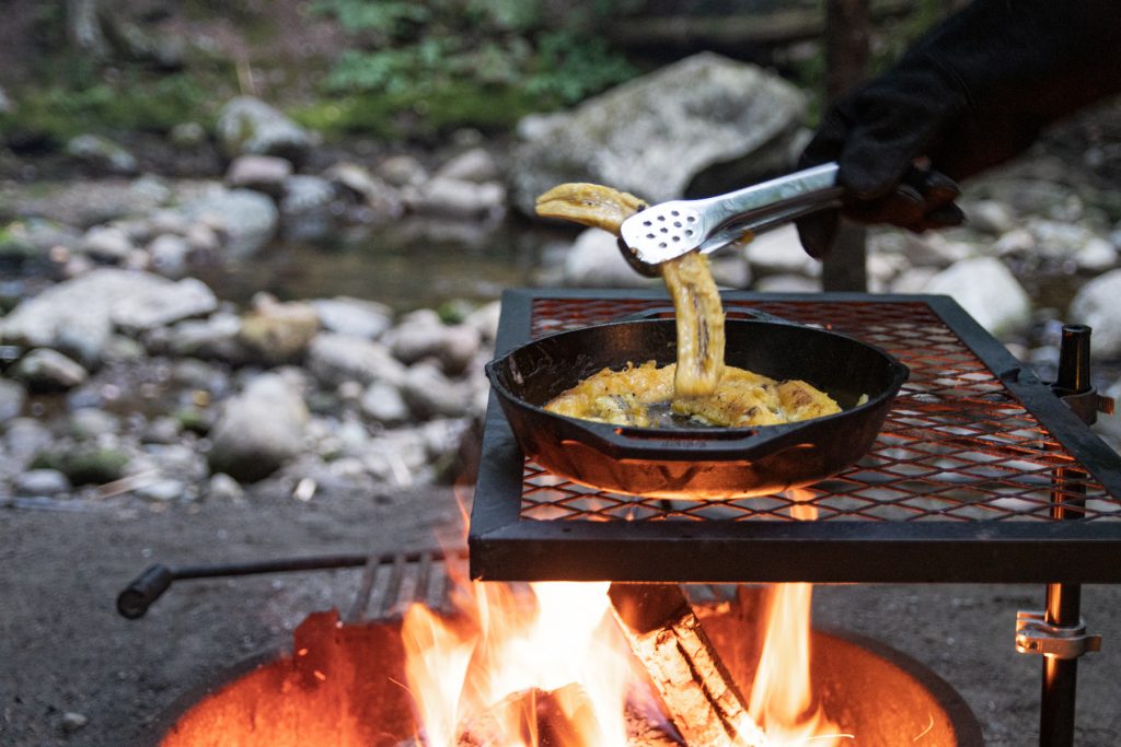 Wearing fire-resistant cloves, a person flips over plantains frying in coconut oil in a Lodge cast iron pan and on top of a campfire grate with a campfire roaring below. A stoney brook can be seen in the back.