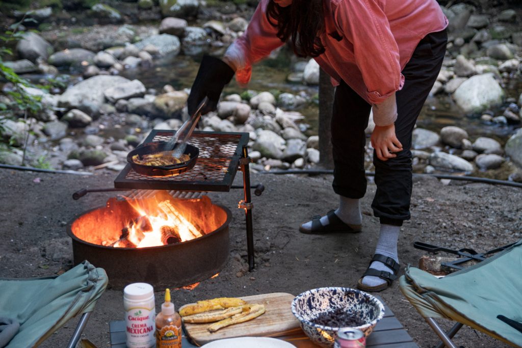 With a stoney creek behind, a woman carefully flips the frying plantains over with tongs.