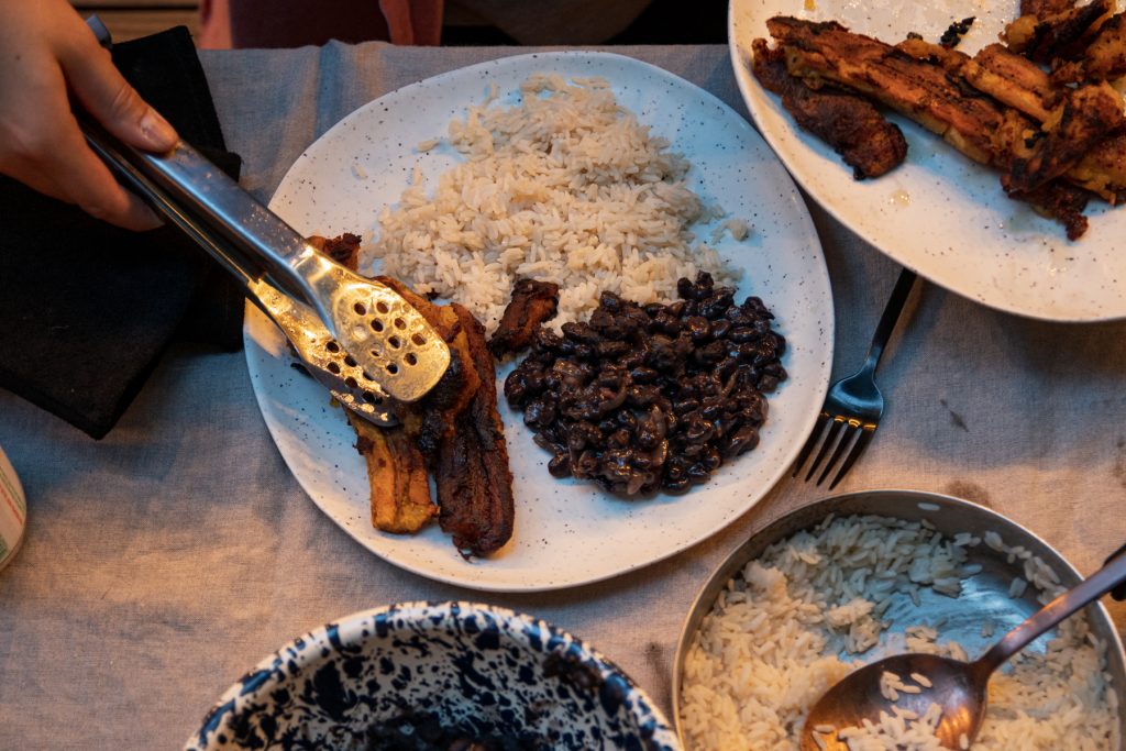 A person serves fried plantains onto a plate with black beans and rice.