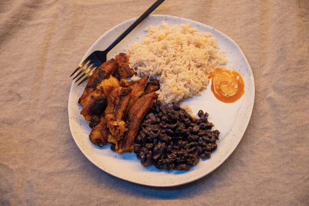 A top-down image of a plate full of fried plantains, rice, black beans, crema Mexicana, and hotsauce