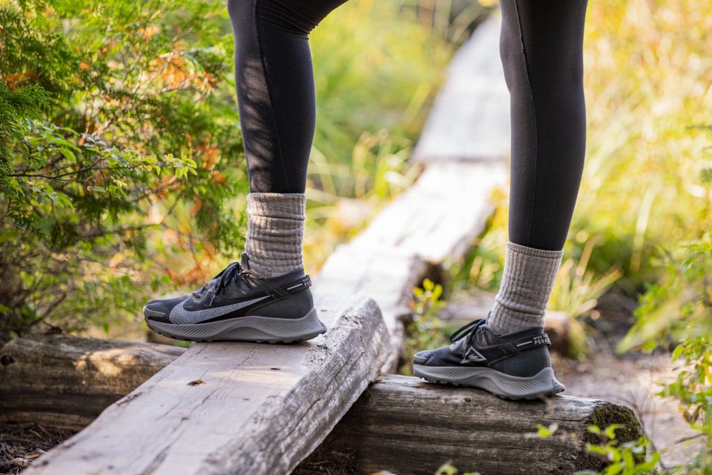 A woman wearing black leggings standing on a log wearing black Nike trail running tennis shoes.