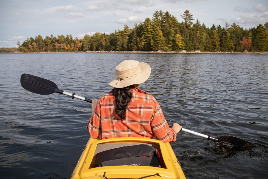 A woman in LL Bean's classic flannel shirt that is red and plaid, kayaking in a yellow kayak. 