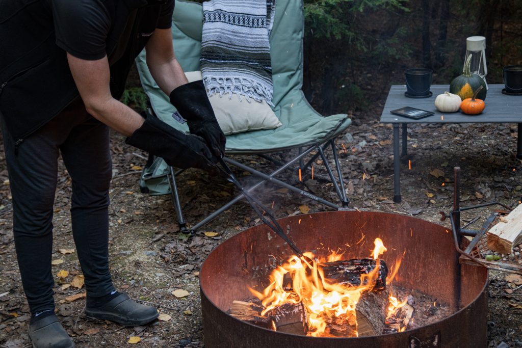 A man wearing heat-resistant gloves and using the log grabber while above a roaring campfire flame.