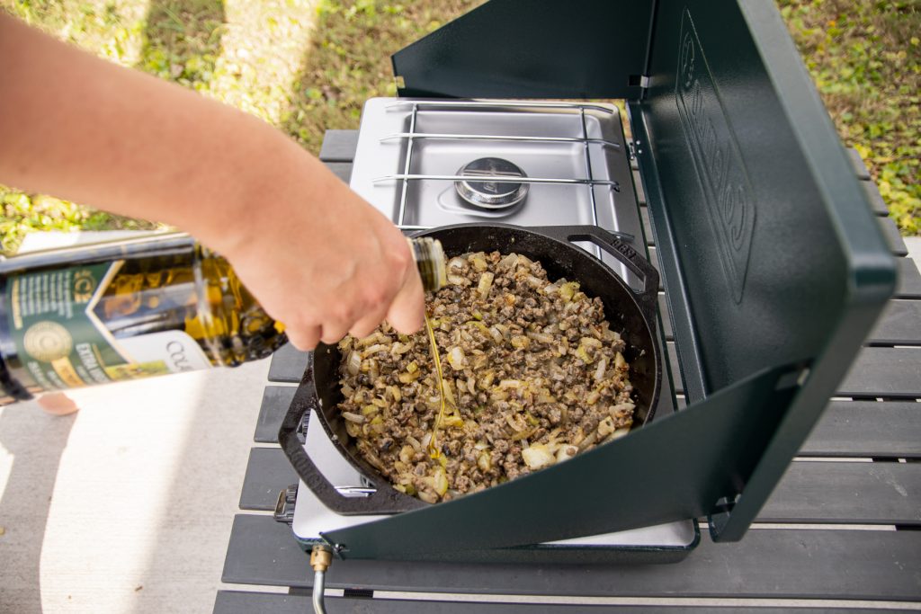 A person pouring olive oil into a Lodge Cast Iron double-handle pan that is filled with Beyond Meat ground beef, all on top of a Coleman portable propane glass classic stove.