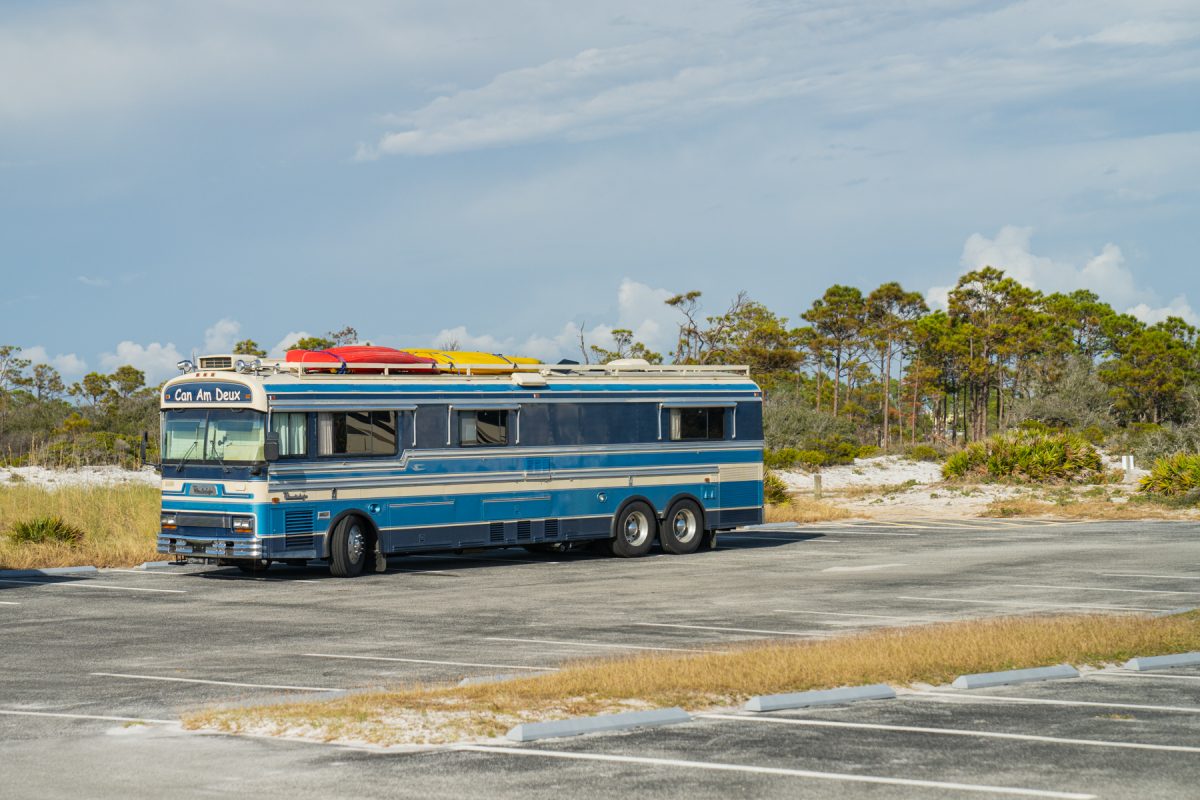 A blue motorhome RV with canoes tied to the top sits in a parking lot surrounded by dunes.