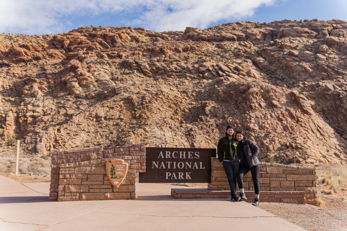 A man and a woman pose in front of the Arches National Park sign in Moab, Utah. Utah's famous red rock can be seen in the background.