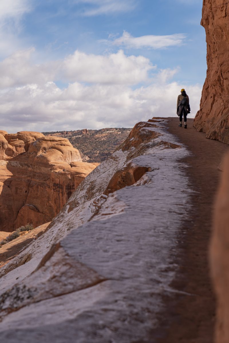 A hiker walks along the rock edge toward the end of the Delicate Arch trail in Arches National Park, Utah. Snow covers the edges of the trail.