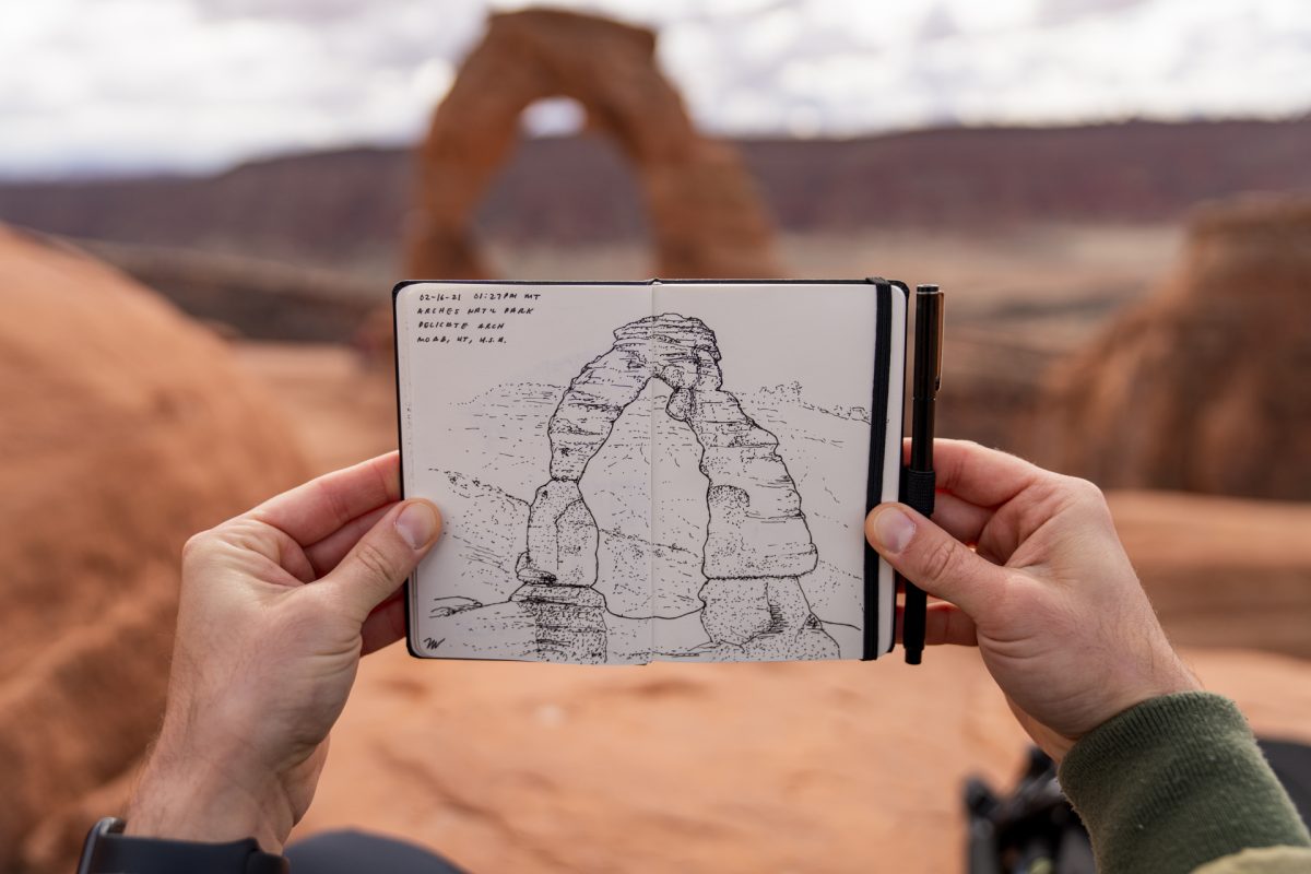 A man holds up his finished drawing of Delicate Arch with the arch in the background at Arches National Park in Moab, Utah.