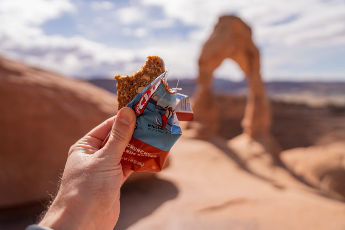 A man holds up a Clif bar while looking out to Delicate Arch in Arches National Park in Moab, Utah.