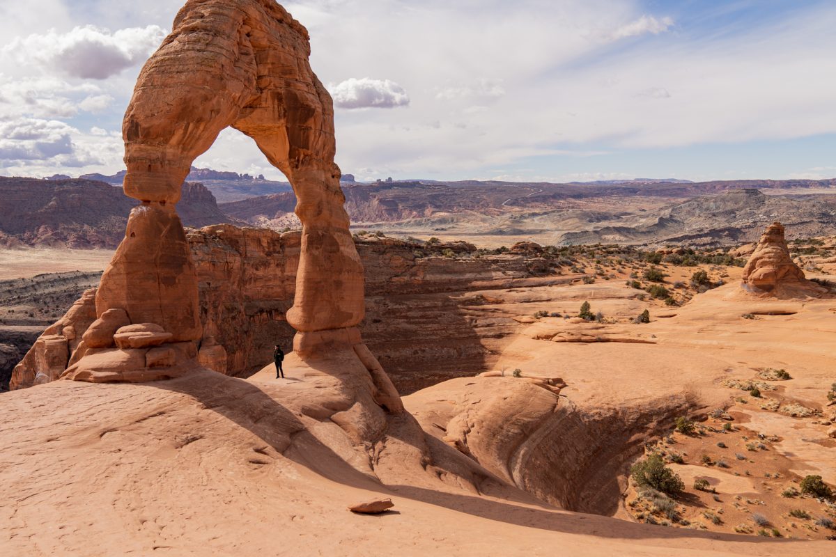 A man stands underneath Delicate Arch with vast desert and rock formations seen in the background at Arches National Park in Moab, Utah.