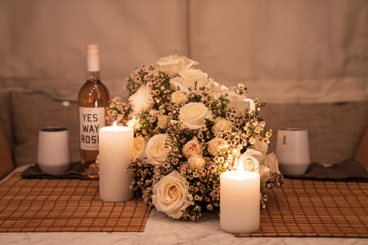 A floral arrangement of roses and wax flowers sits on an RV dinette table surrounded by lit candles, wine, and Miir wine tumblers.