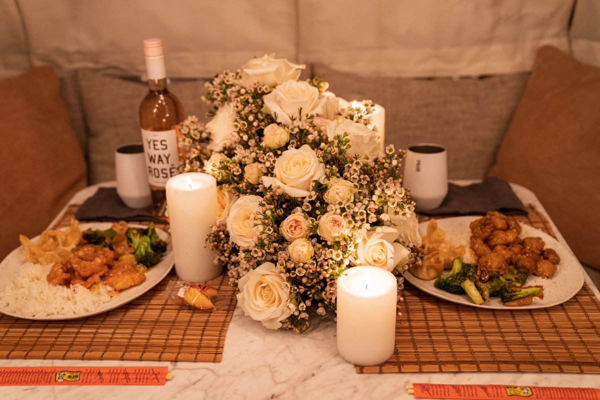 A table spread of Chinese takeout, wine, Miir wine tumblers, lit candles, and a floral arrangement of wax flowers and roses.