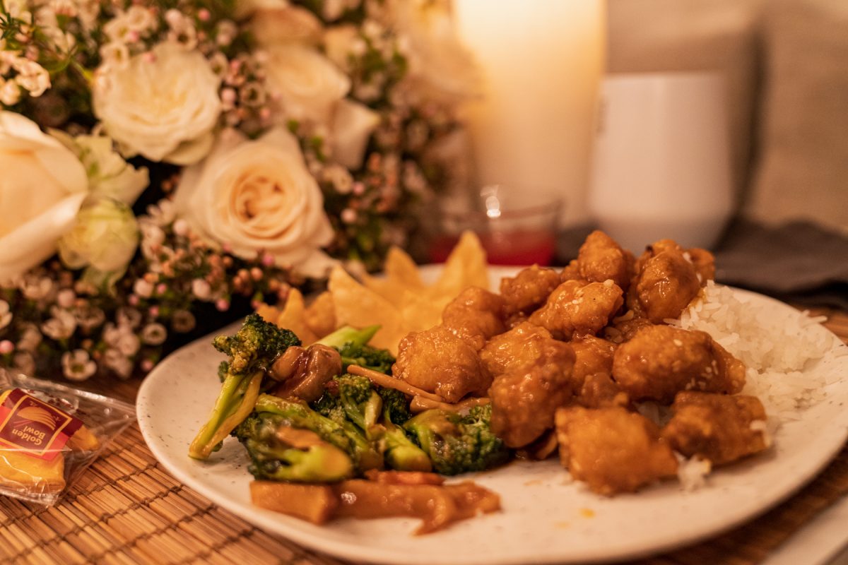 A plate of Chinese takeout, consisting of sesame chicken, vegetable stir fry, and crab rangoon with white rice. In the background is a fortune cookie, a floral arrangement of roses and wax flowers, candles, and a wine tumbler.