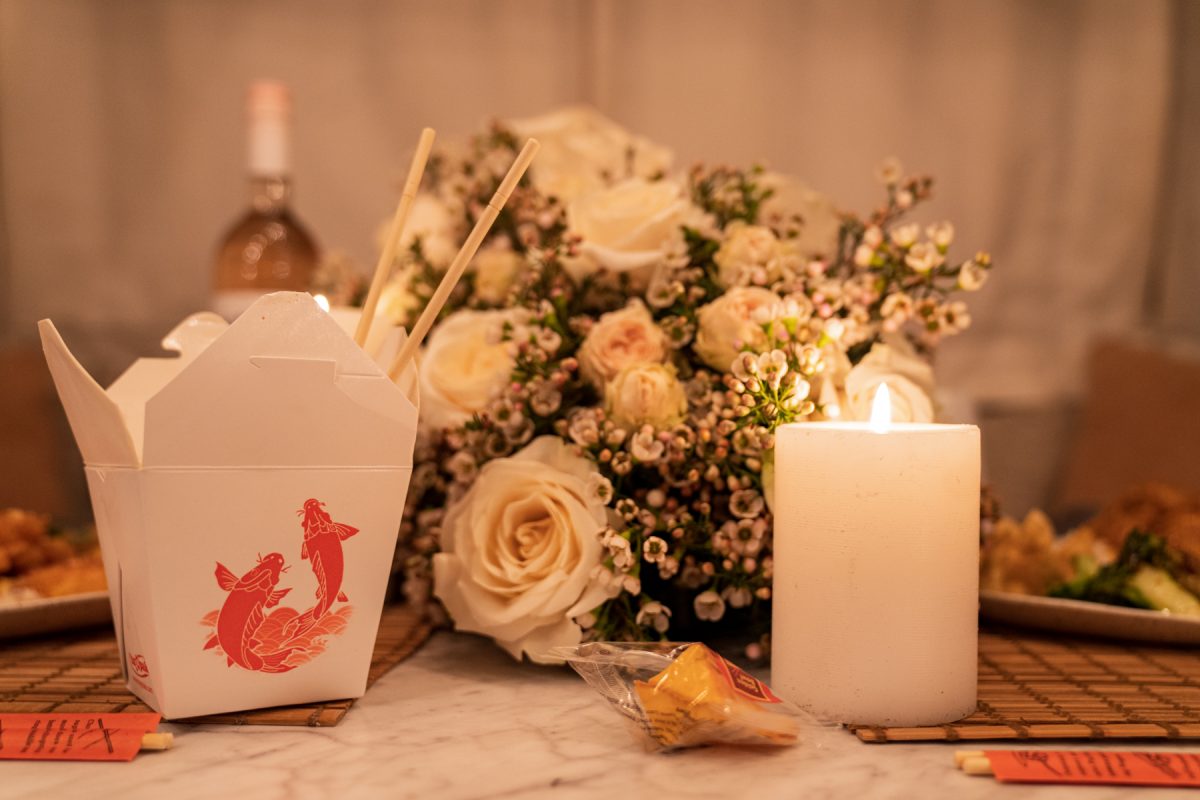 A Chinese takeout box sits next to a lit candle and a floral arrangement of roses and wax flowers. A fortune cookie sits nearby.