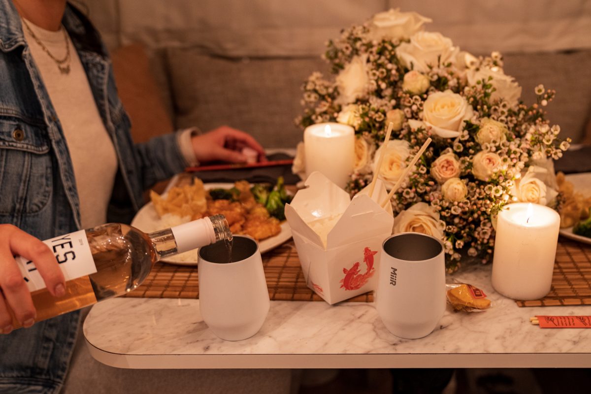 A woman pours rosé wine into a white Miir wine tumbler that is sitting next to her plate of Chinese takeout.