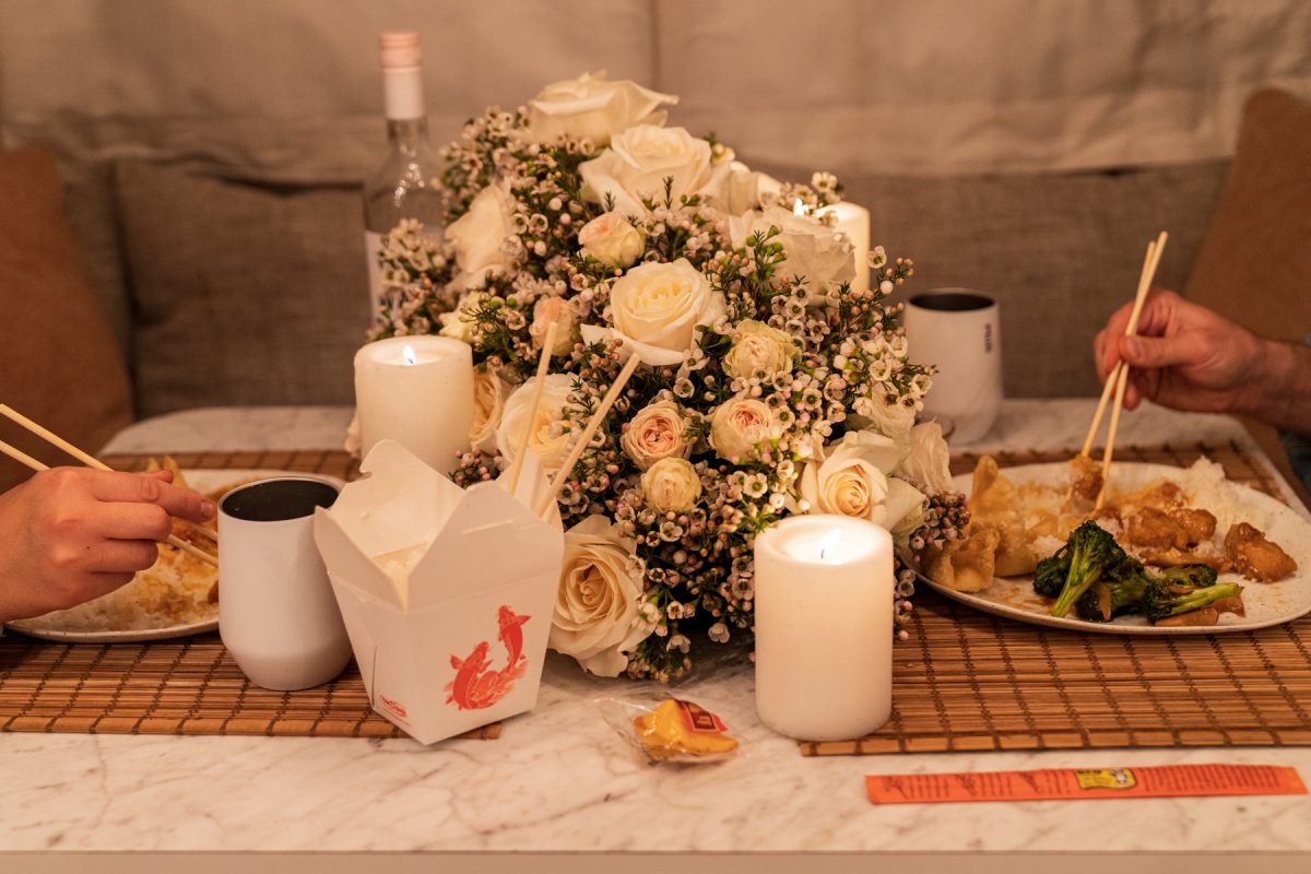 A couple enjoys Chinese takeout while celebrating Valentine's Day by having date night inside their Airstream RV trailer. The pair are using chopsticks. Candles are lit and a floral arrangement of roses and wax flowers sit in the middle of the table.