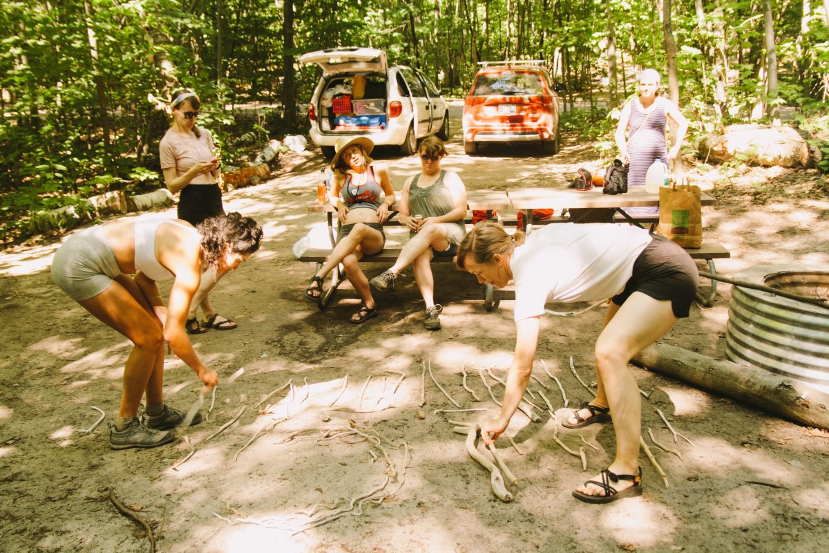 A group of six women at a campsite, some of them sitting on the picnic table, others playing with sticks on the ground.