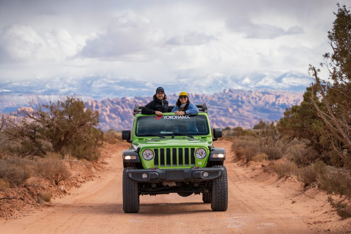 A couple peeks their heads out of the sunroof of a green jeep on the Gemini Bridges OHV trail in Moab Utah. The La Sal Mountains are in the back.