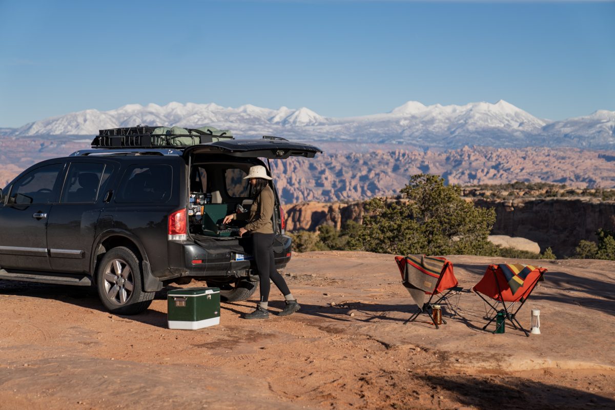 A woman makes grilled cheese on a camp stove in the back of a black SUV. In the background, the La Sal Mountains near Moab, Utah, can be seen. 