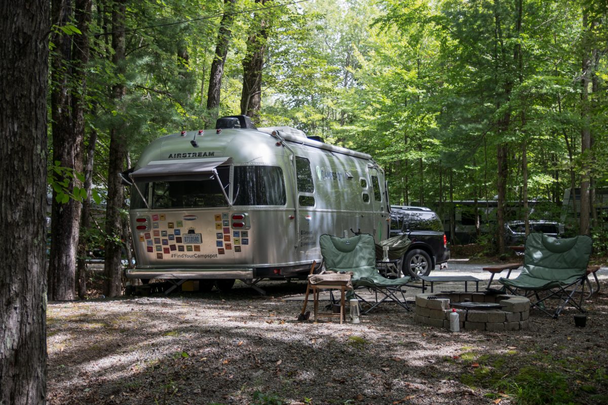 An Airstream trailer with a bunch of bumper stickers sits in a wooded area with two camper chairs in front of it.