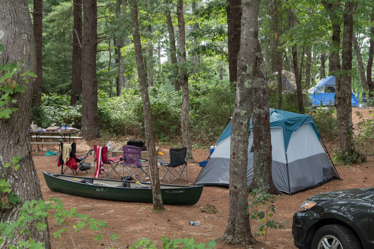 A tent sits at a campsite with a canoe and camping chairs nearby. The campsite is surrounded by trees.