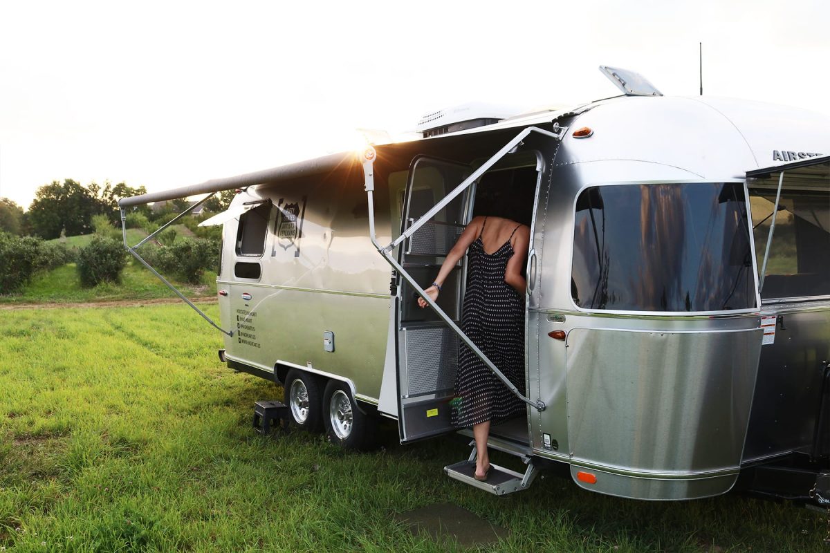 A woman in a navy dress walks into her Airstream trailer with the sun setting above the awning of the trailer.