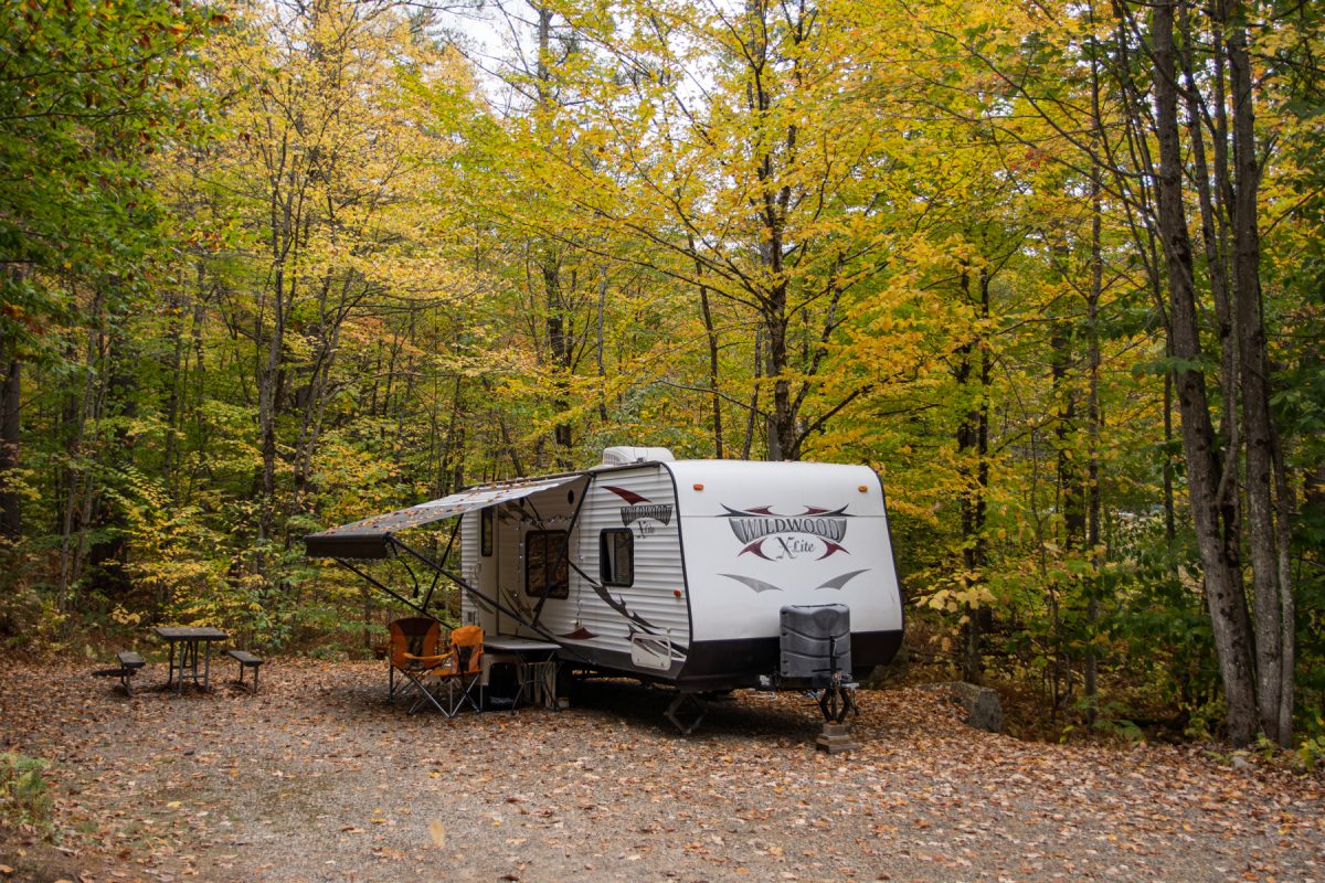 An RV sits at a campsite covered in fall leaves with orange camp chairs underneath.