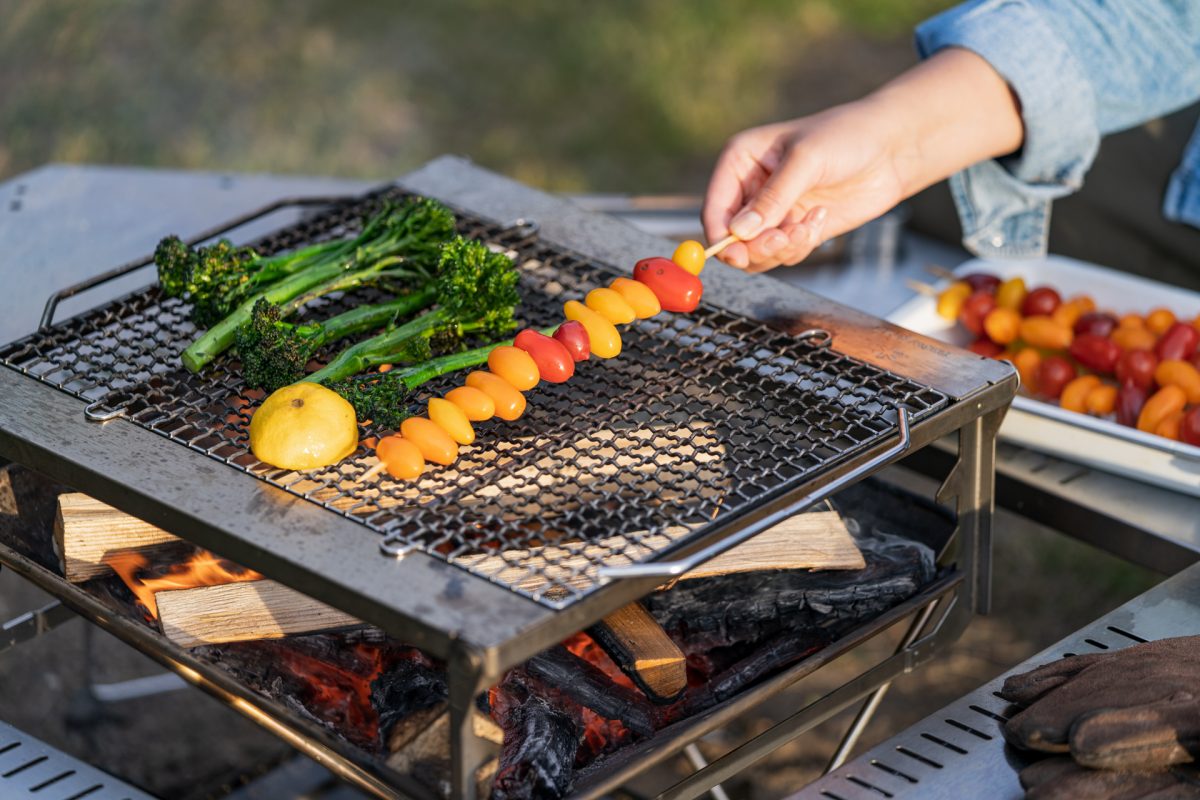 A person places skewered tomatoes and broccolini onto a campfire grate while making campfire roasted tomato pasta.