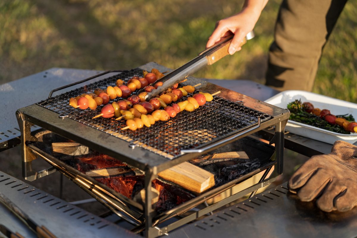 A person flips over skewered tomatoes on a campfire grill while making campfire roasted tomato pasta with burrata and broccolini. 