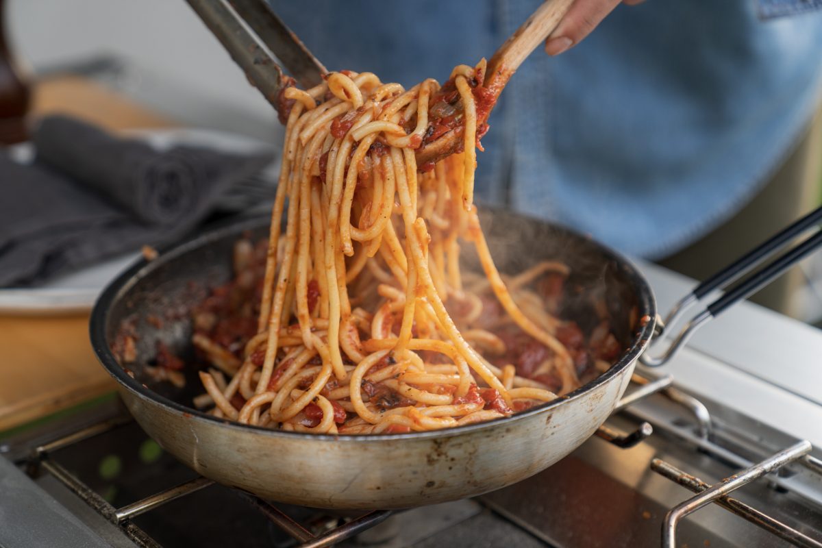 A person tosses pasta in the roasted tomato pasta sauce while making campfire roasted tomato pasta with broccolini and burrata.