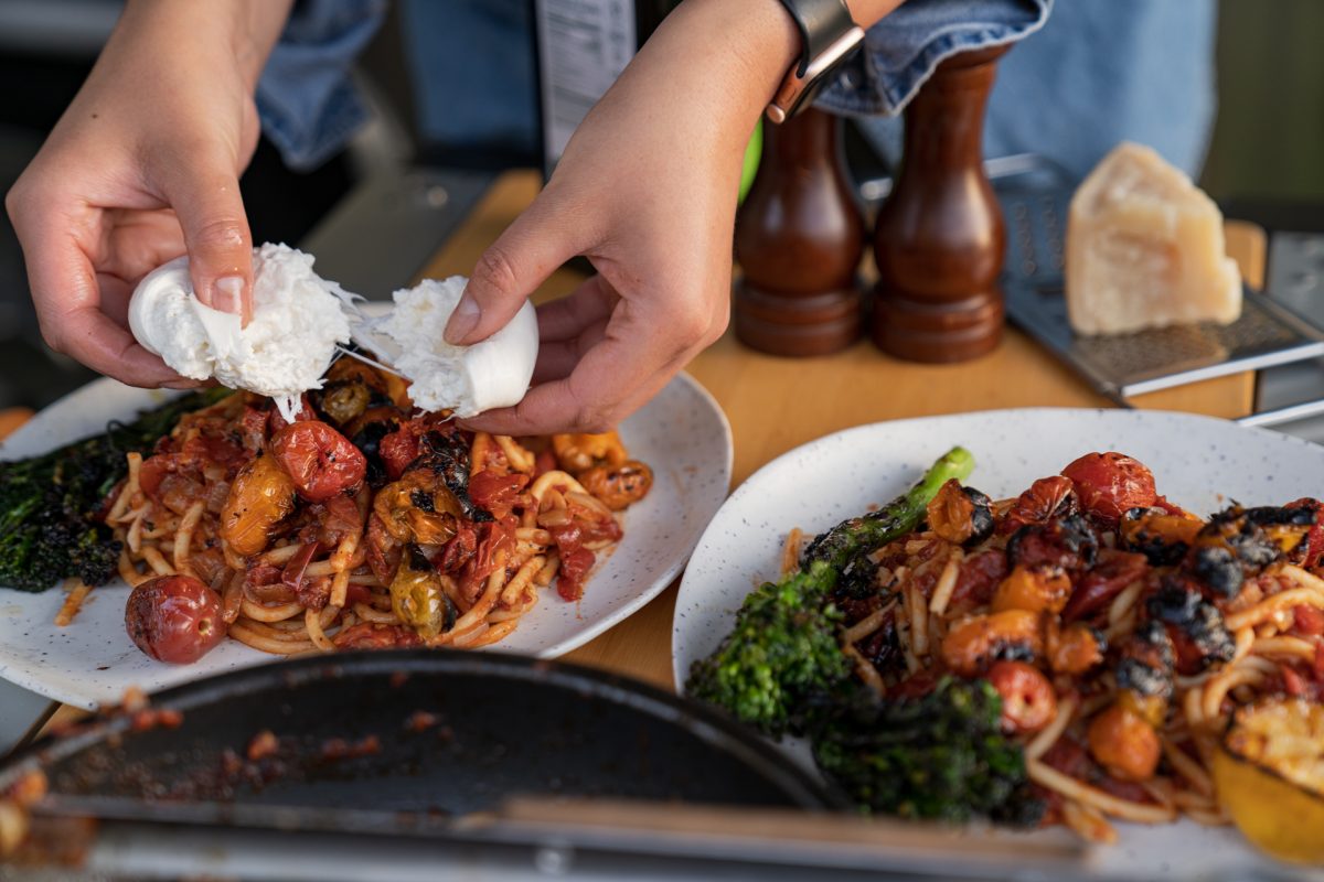 Someone splits a ball of burrata onto a plate of campfire roasted tomato pasta. 