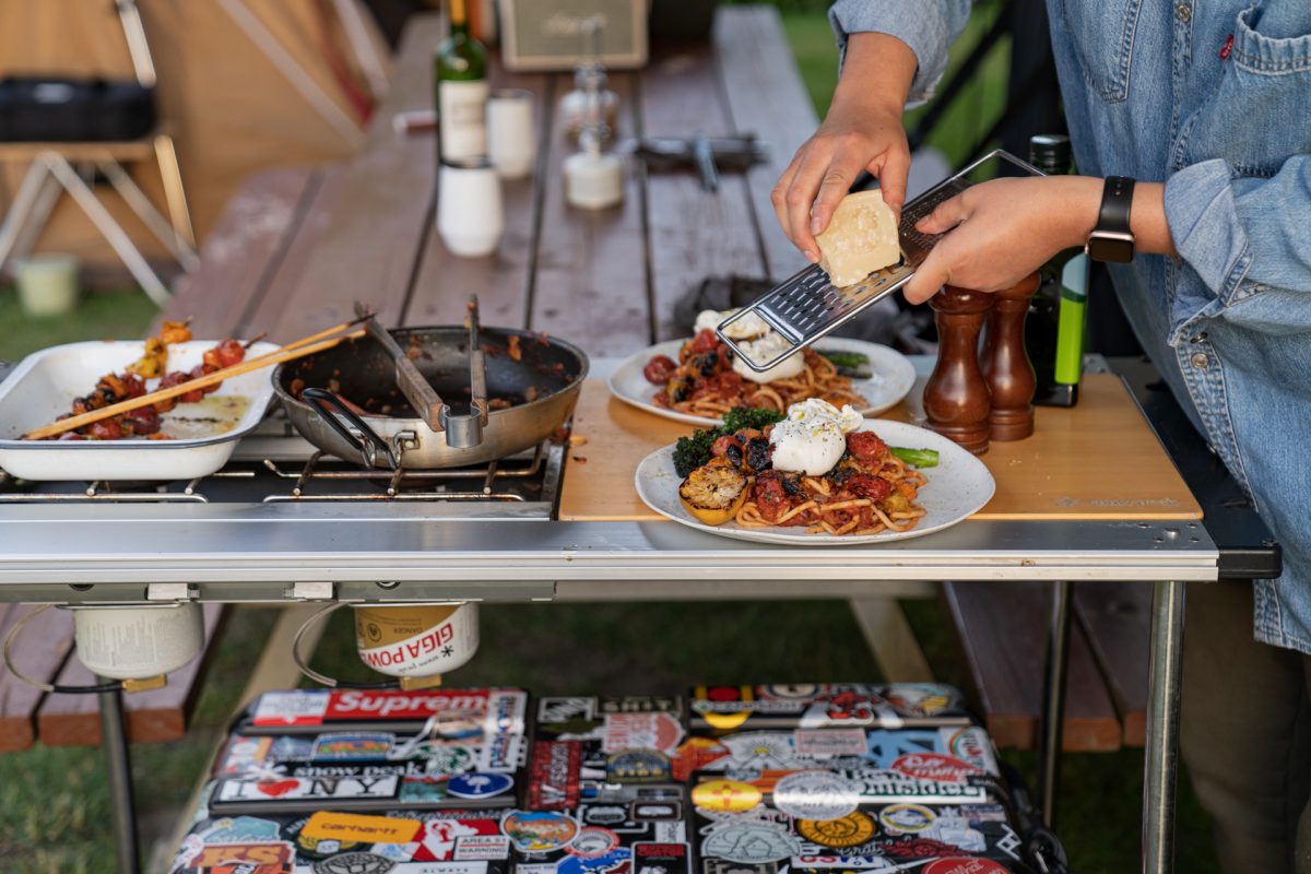 A camper grates parmesan cheese onto a plate of campfire roasted tomato pasta with burrata and broccolini. 