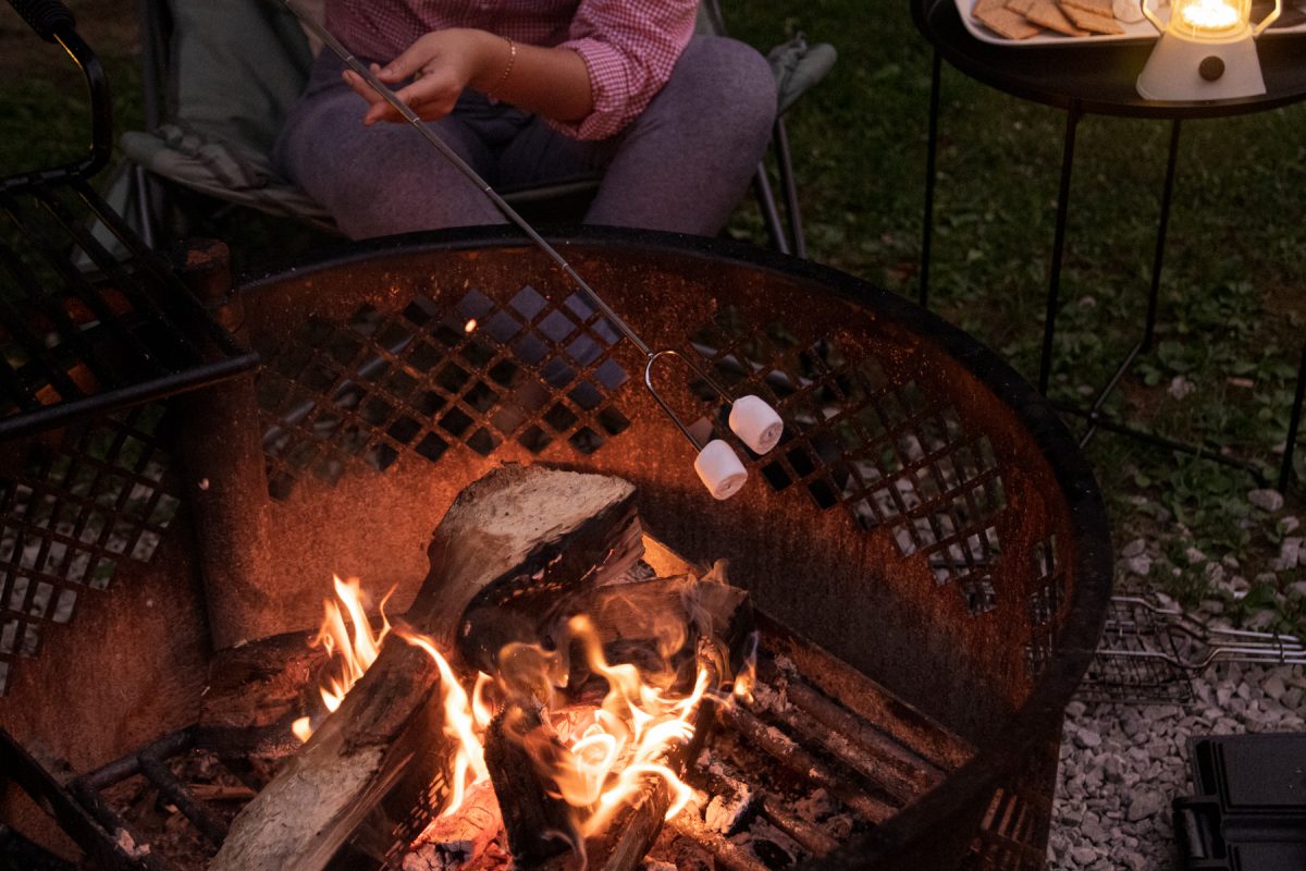 A person roasts their marshmallows on top of a campfire with a lantern glowing in the back.