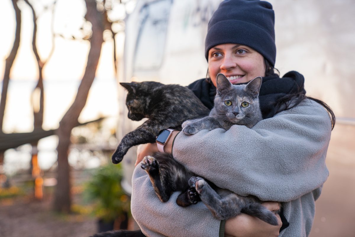 A woman holds two cats up, smiling with a hammock and campsite in the background.